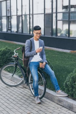 smartphone kullanarak ve bisiklet üzerinde oturan kağıt bardak ile gülümseyen Asyalı adam