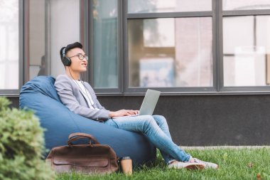 Asya fasulye çanta sandalyede otururken dizüstü bilgisayar ve bir kulaklık ile müzik dinleme adamım