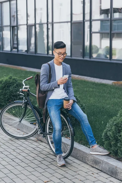 Sonriente Asiático Hombre Con Papel Taza Usando Smartphone Sentado Bicicleta — Foto de stock gratuita