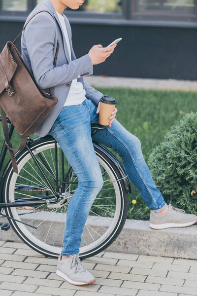 cropped view of man with coffee to go using smartphone and sitting on bike