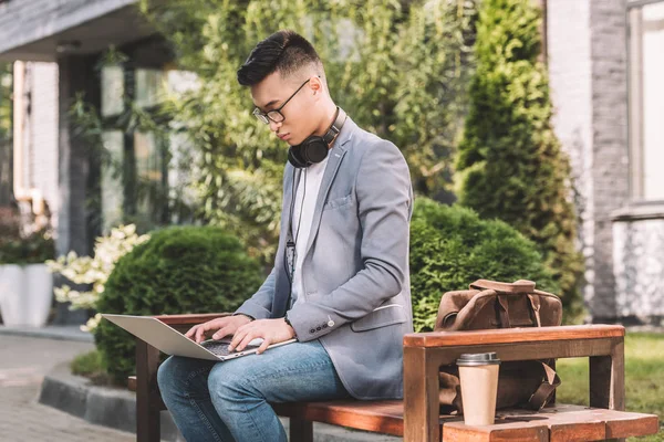 asian teleworker using laptop while sitting on bench with leather bag and coffee to go