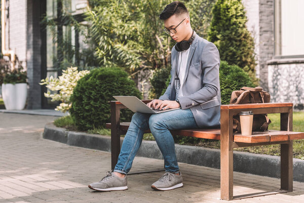 confident asian freelancer working on laptop while sitting on bench with leather bag and coffee to go