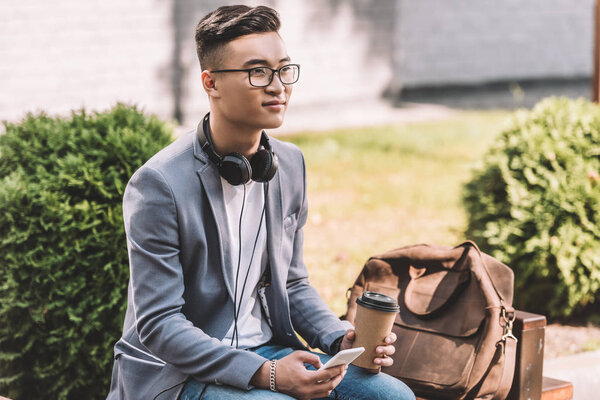 asian man with coffee to go, smartphone and headphones sitting on bench with leather backpack