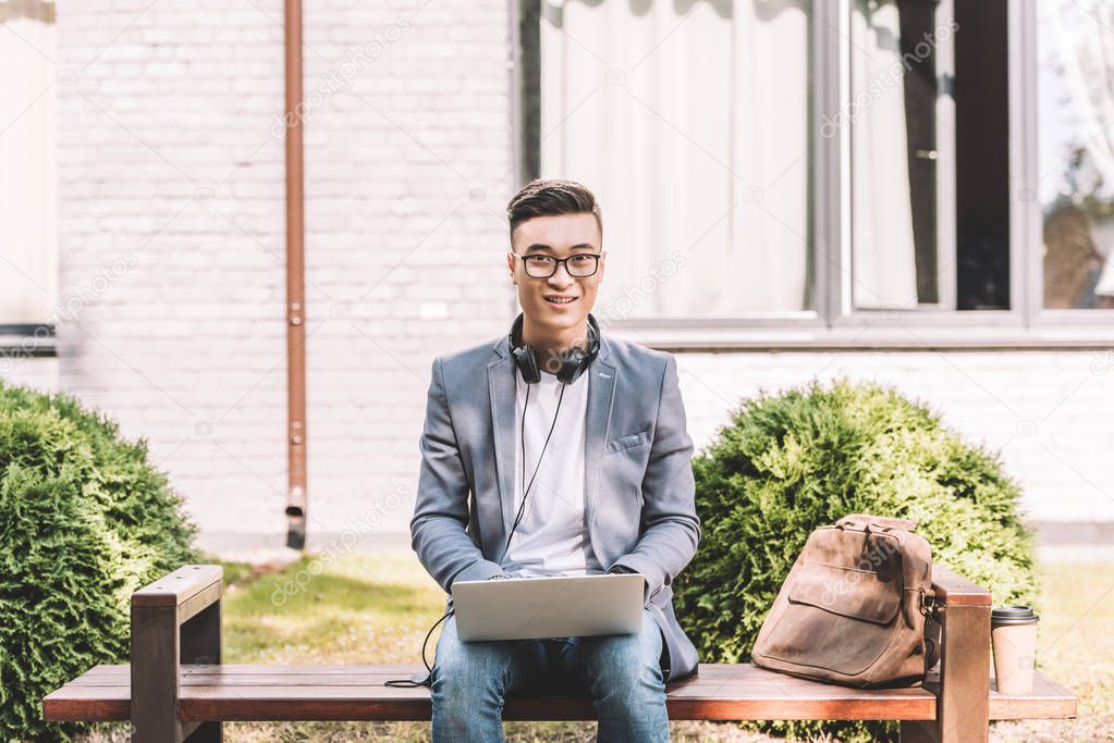 smiling asian freelancer working on laptop while sitting on bench with leather bag