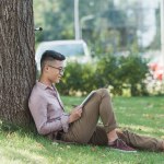 Vista lateral do homem asiático em óculos usando tablet digital na grama verde no parque