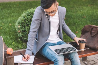 Asya freelancer bankta smartphone ve dizüstü bilgisayar kullanırken planlayıcısı olarak yazma