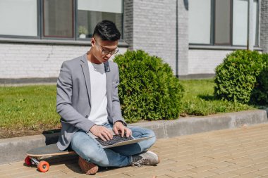 Asya adam sokakta longboard üzerinde otururken dizüstü bilgisayarda yazarak