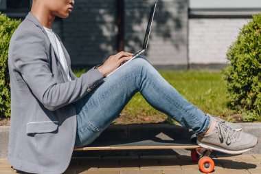 partial view of teleworker using laptop while sitting on longboard on street clipart