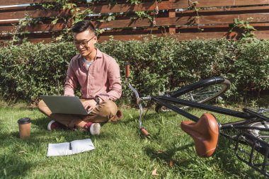 Asya freelancer laptop bisiklet ve park içinde defter ile yeşil çimenlerin üzerinde çalışma