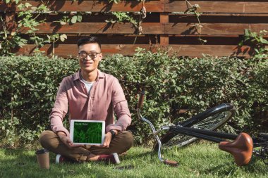 yeşil çimenlerin üzerinde oturan ve tablet online ticaret yazıt ile parkta elinde ekranda gösteren Asyalı adam gülümseyen