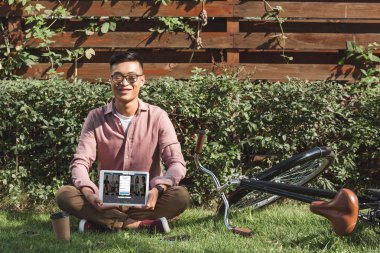yeşil çimenlerin üzerinde oturan ve tablet LinkedIn logosu ile parkta elinde ekranda gösteren Asyalı adam gülümseyen