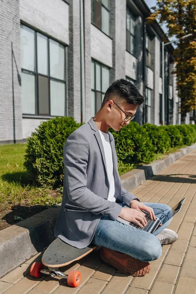 Asian Man Typing Laptop While Sitting Longboard Street — Free Stock Photo