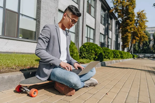 Pekerja Lepas Asia Menggunakan Laptop Ketika Duduk Longboard Jalan — Foto Stok Gratis