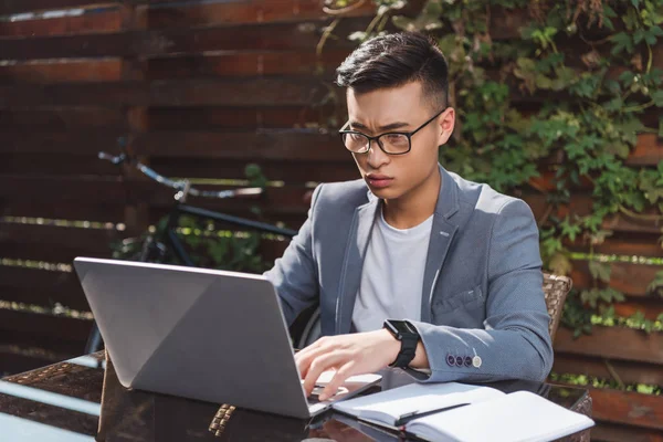 focused asian businessman remote working on laptop at tabletop with notebook in cafe