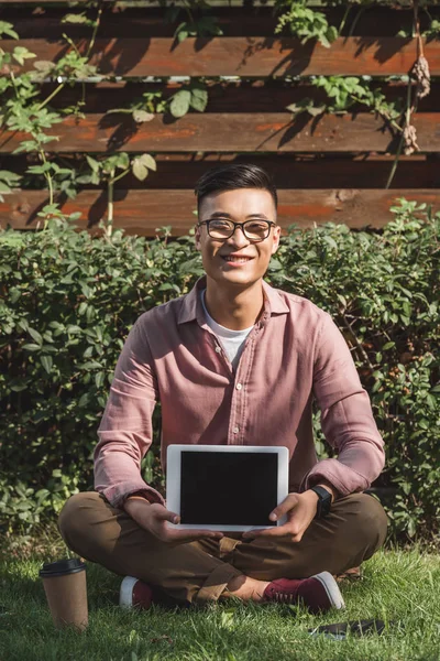 Lächelnder Asiatischer Mann Sitzt Auf Grünem Gras Und Zeigt Tablet — kostenloses Stockfoto