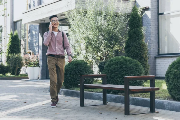 asian man with backpack having conversation on smartphone while walking on street