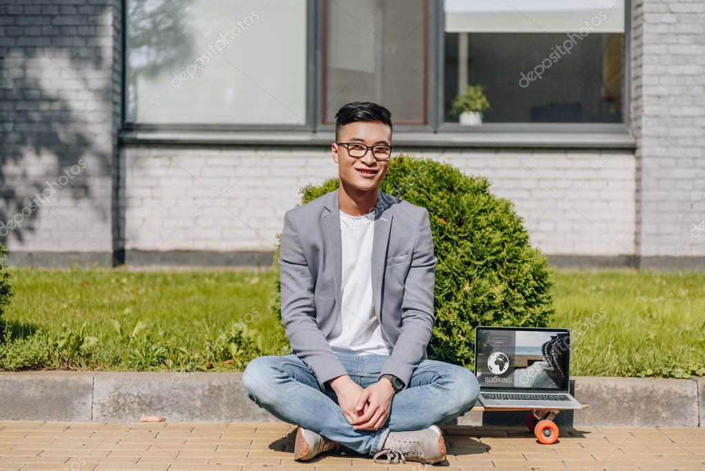 Smiling asian man sitting on longboard with laptop with booking website on screen on street