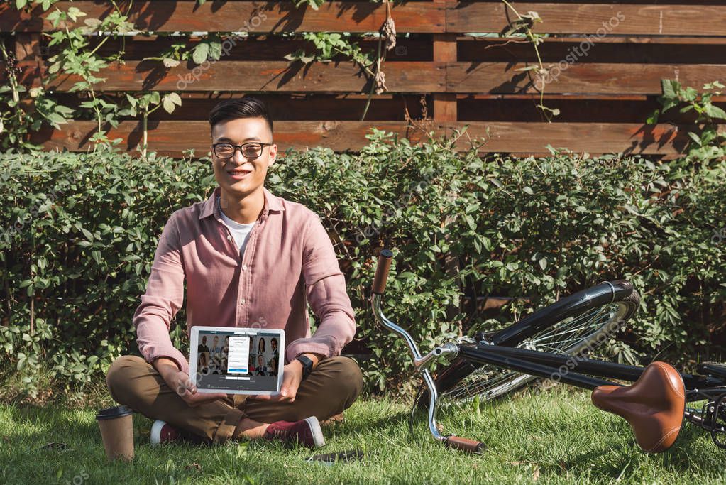Smiling asian man sitting on green grass and showing tablet with linkedin logo on screen in hands in park