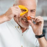 Guapo sonriente hombre divertirse con verduras durante la preparación de ensalada para la cena en casa