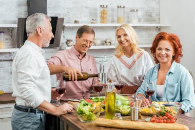 handsome man pouring wine to happy old friends during dinner in kitchen