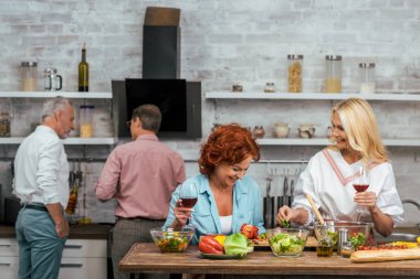 laughing beautiful women preparing salad for dinner and holding wineglasses at home