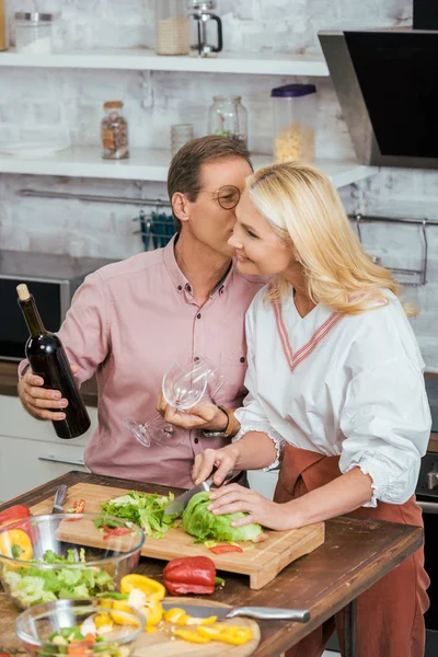 husband kissing wife and holding bottle of wine during dinner at home