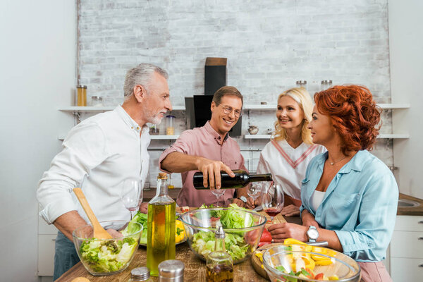 handsome smiling man pouring wine to happy old friends during dinner in kitchen