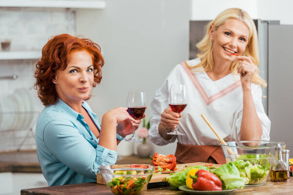 smiling attractive women preparing salad for dinner, holding wineglasses and looking at camera at home