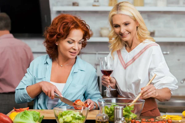 Smilende Attraktive Kvinder Forbereder Salat Til Middag Med Vin Derhjemme - Stock-foto