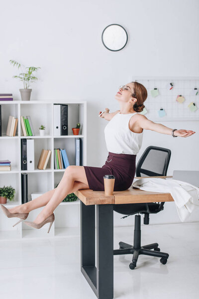 счастливая деловая женщина с протянутыми руками сидит на столе во время кофе-брейка в современном офисе
