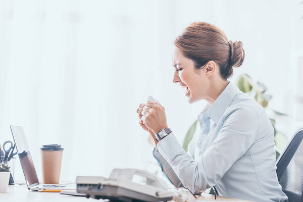 Взгляд сбоку на сумасшедшую взрослую предпринимательницу, сжимающую бумагу на рабочем месте
