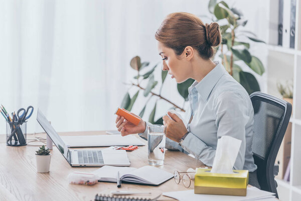 side view of sick adult businesswoman sitting at workplace with plastic jar of pills and water