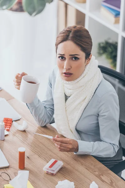 high angle view of sick businesswoman in scarf holding cup of tea and electric thermometer sitting at office and looking at camera