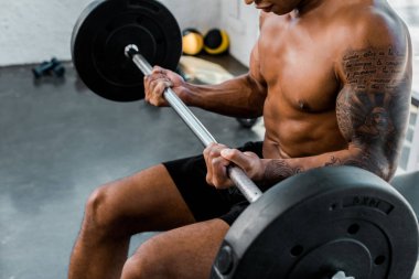 cropped shot of athletic young african american sportsman sitting on bench and lifting barbell in gym clipart