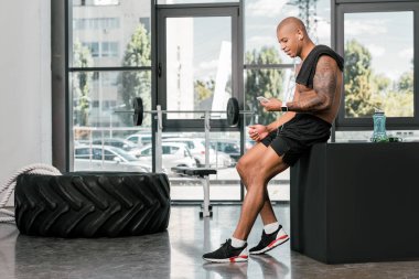 oturma ve spor salonunda smartphone kullanarak kulaklık gülümseyen genç sporcu yan görünüm