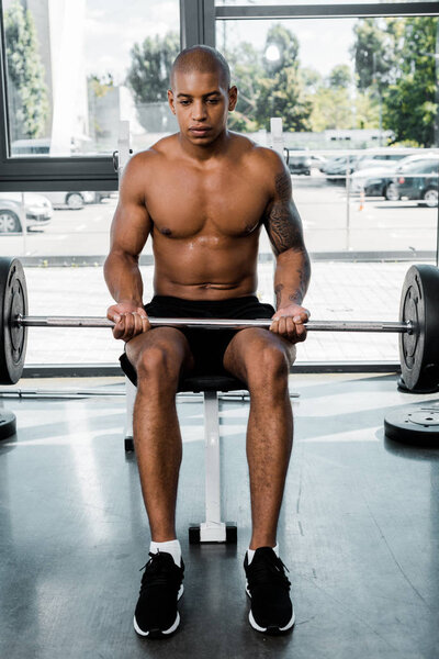 athletic young african american sportsman sitting on bench and lifting barbell in gym