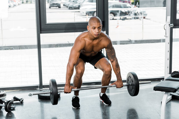 Молодой африканский спортсмен без рубашки поднимает штангу в спортзале
