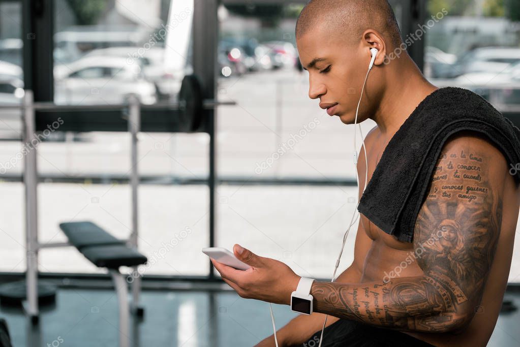 side view of muscular shirtless young man in earphones sitting and using smartphone in gym