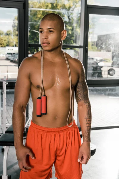 young shirtless african american athlete standing with jump rope over neck at gym