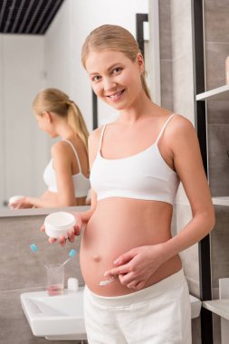 happy pregnant woman applying cream on tummy to avoid stretch marks and looking at camera in bathroom clipart