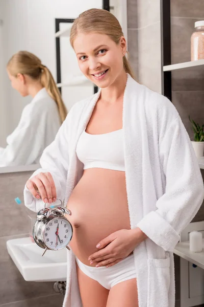 Happy Pregnant Woman Holding Alarm Clock Tummy Looking Camera Bathroom — Free Stock Photo