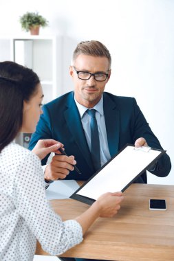 handsome lawyer in eyeglasses giving clipboard with document and pen to female client clipart
