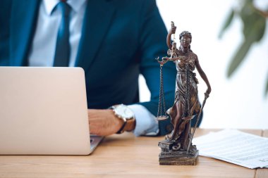 cropped shot of lawyer using laptop and lady justice statue on table clipart