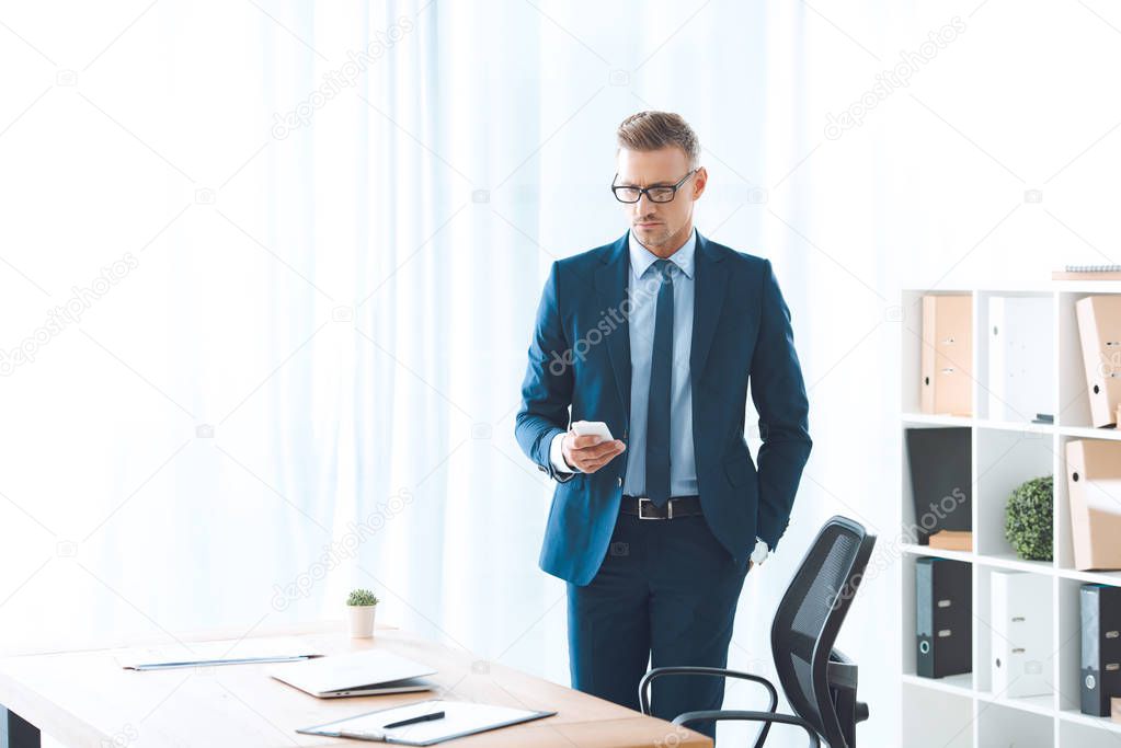 handsome businessman in eyeglasses and formal wear using smartphone at workplace