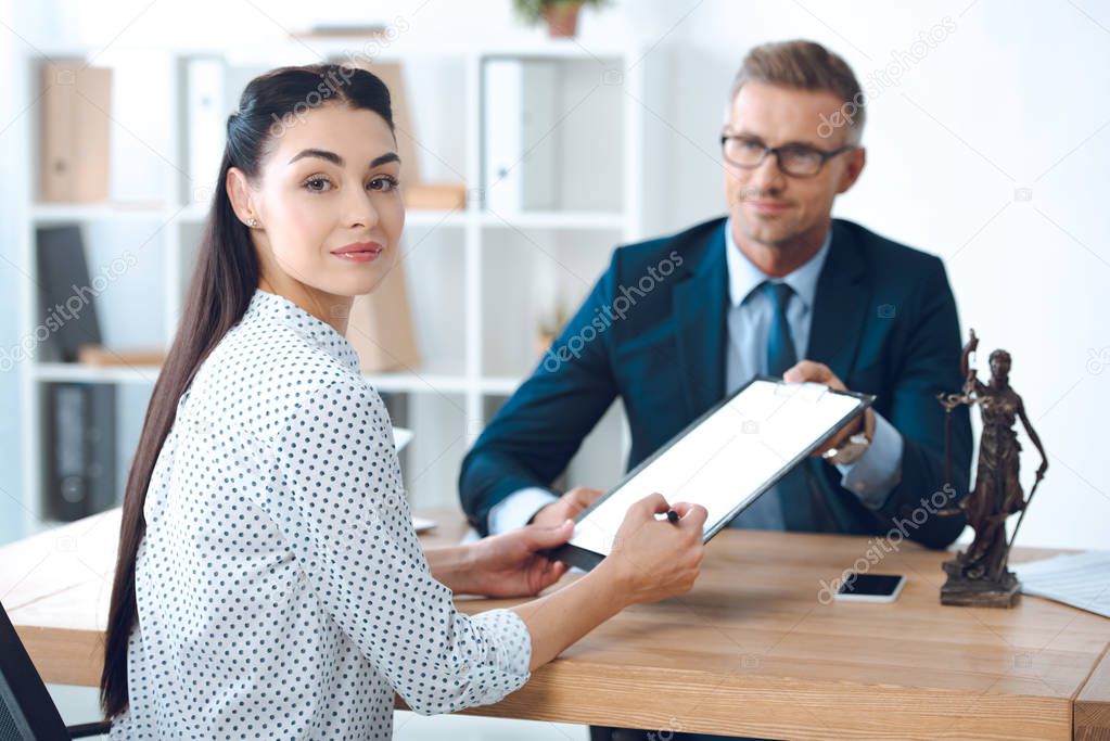 smiling lawyer holding clipboard while young woman signing papers and looking at camera