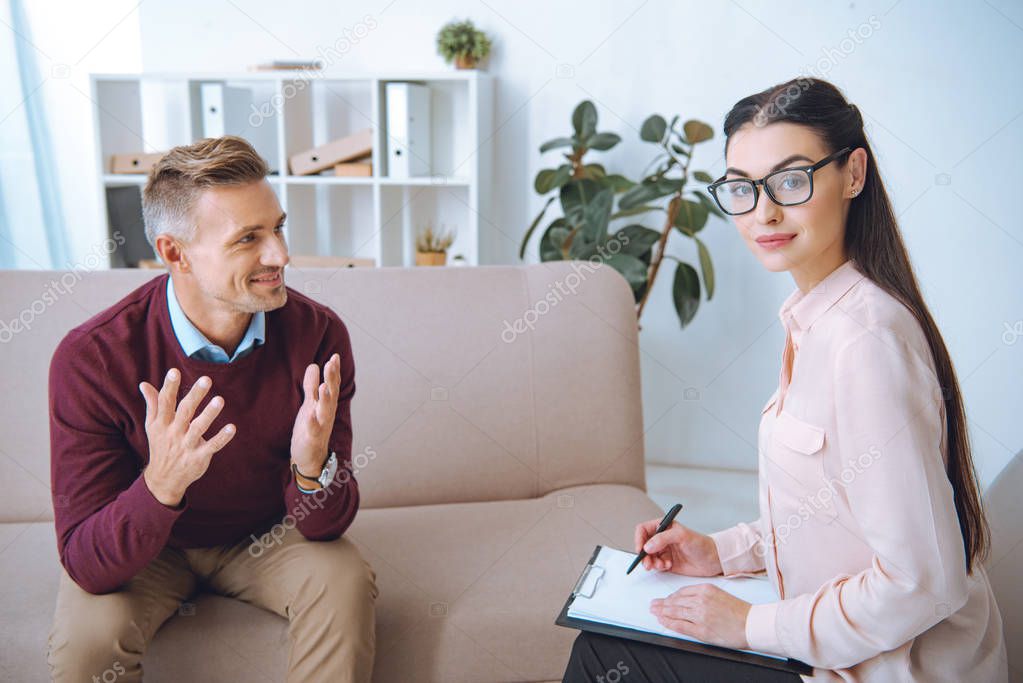 attractive psychologist in eyeglasses taking notes and looking at camera while smiling patient talking in office