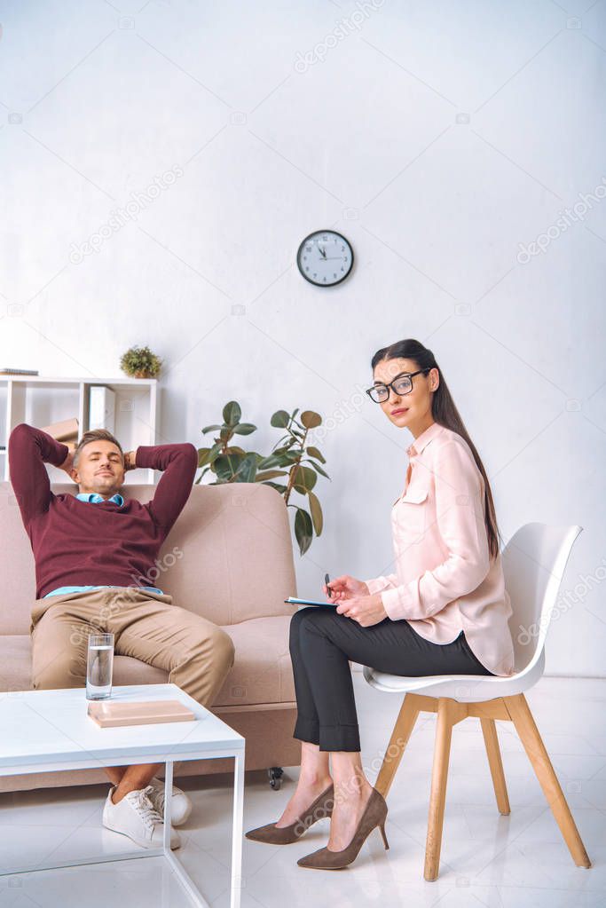 attractive psychotherapist looking at camera while patient resting with hands behind head on couch