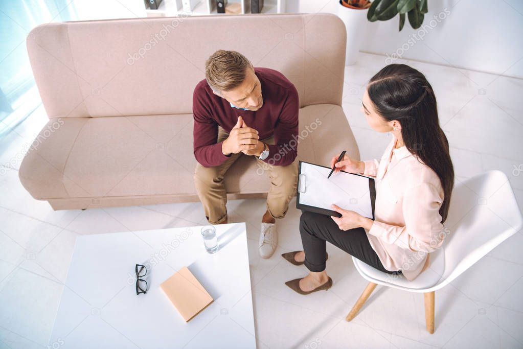 high angle view of psychologist writing on clipboard and looking at patient during therapy