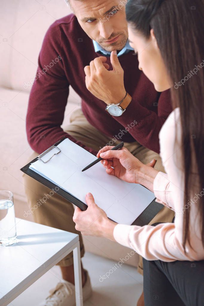female psychologist pointing at blank clipboard during therapy appointment with male patient