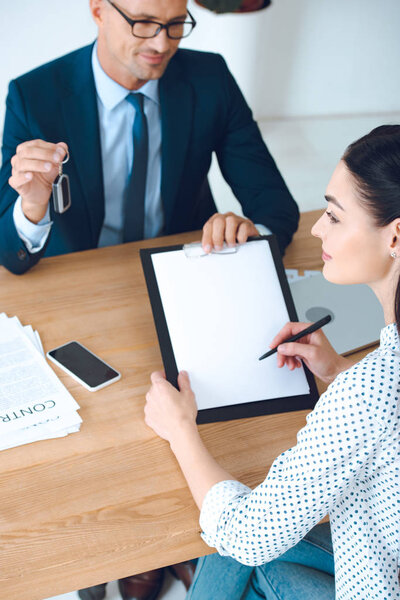high angle view of smiling insurance agent with car key and client signing papers at workplace in office
